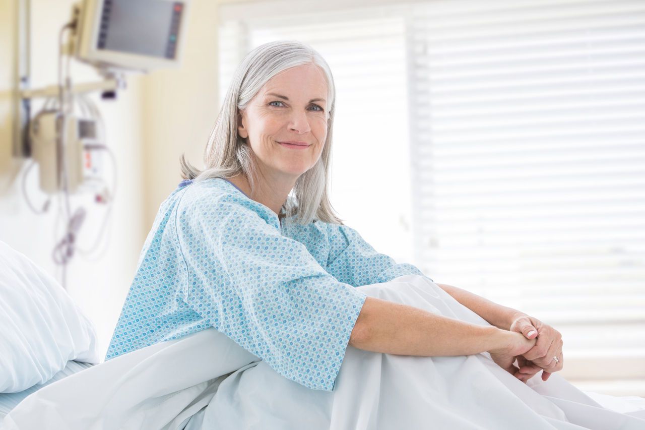 A smiling mature woman in a hospital gown sits on a hospital bed.