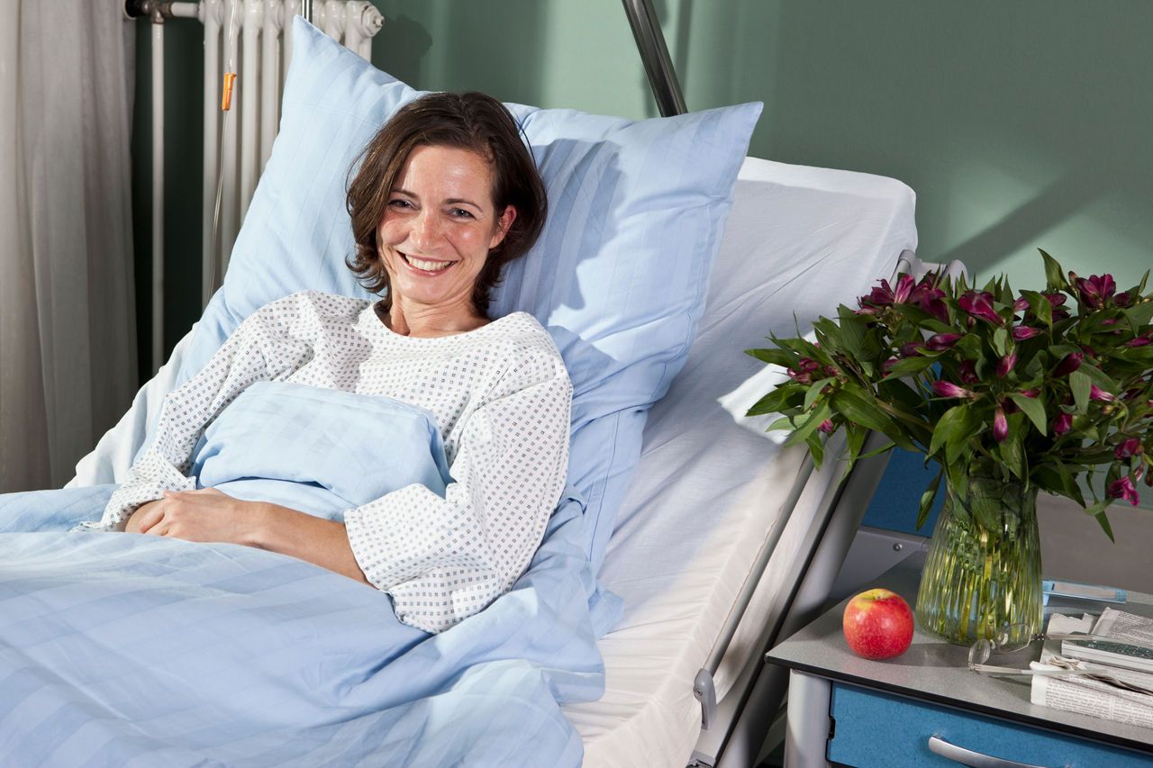 A smiling woman in a hospital bed with flowers and an apple on the nightstand.