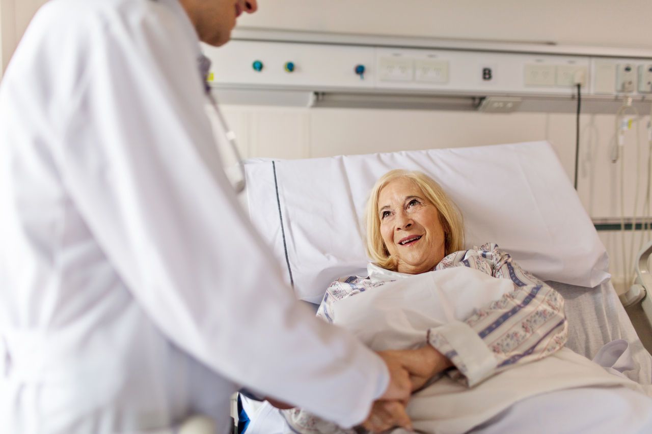 A doctor holds the hand of a smiling elderly woman in a hospital bed.