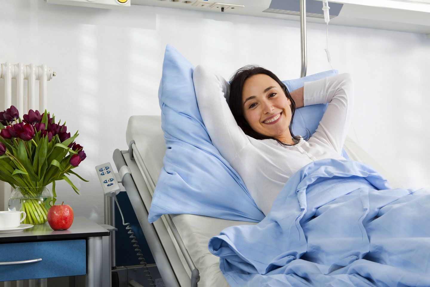 A smiling woman relaxes in a hospital bed with flowers and an apple on the bedside table.